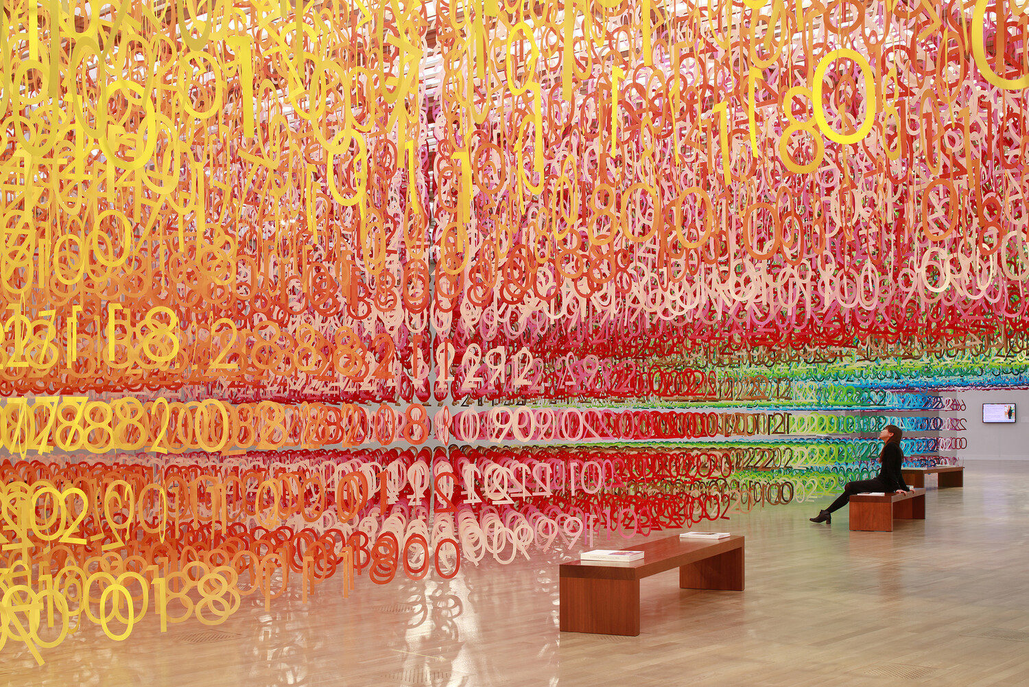 a woman gazes up at multicolored strings of numbers hanging from an art gallery ceiling