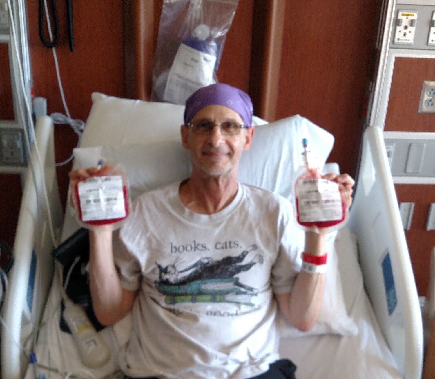 A man sits in a hospital bed and holds up two medical bags with bright red liquid in them