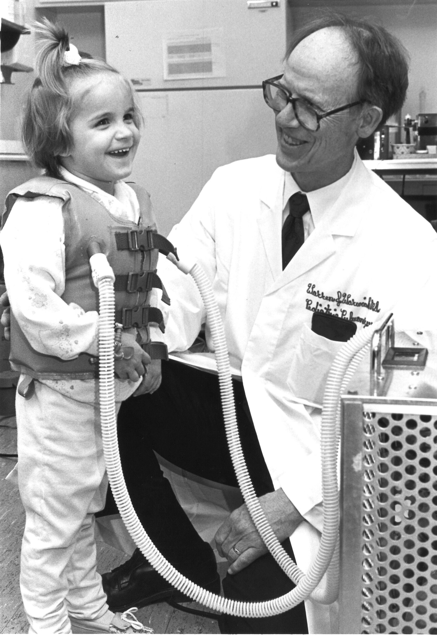 A photo of a young child wearing a chest-compression vest while smiling and standing next to a doctor
